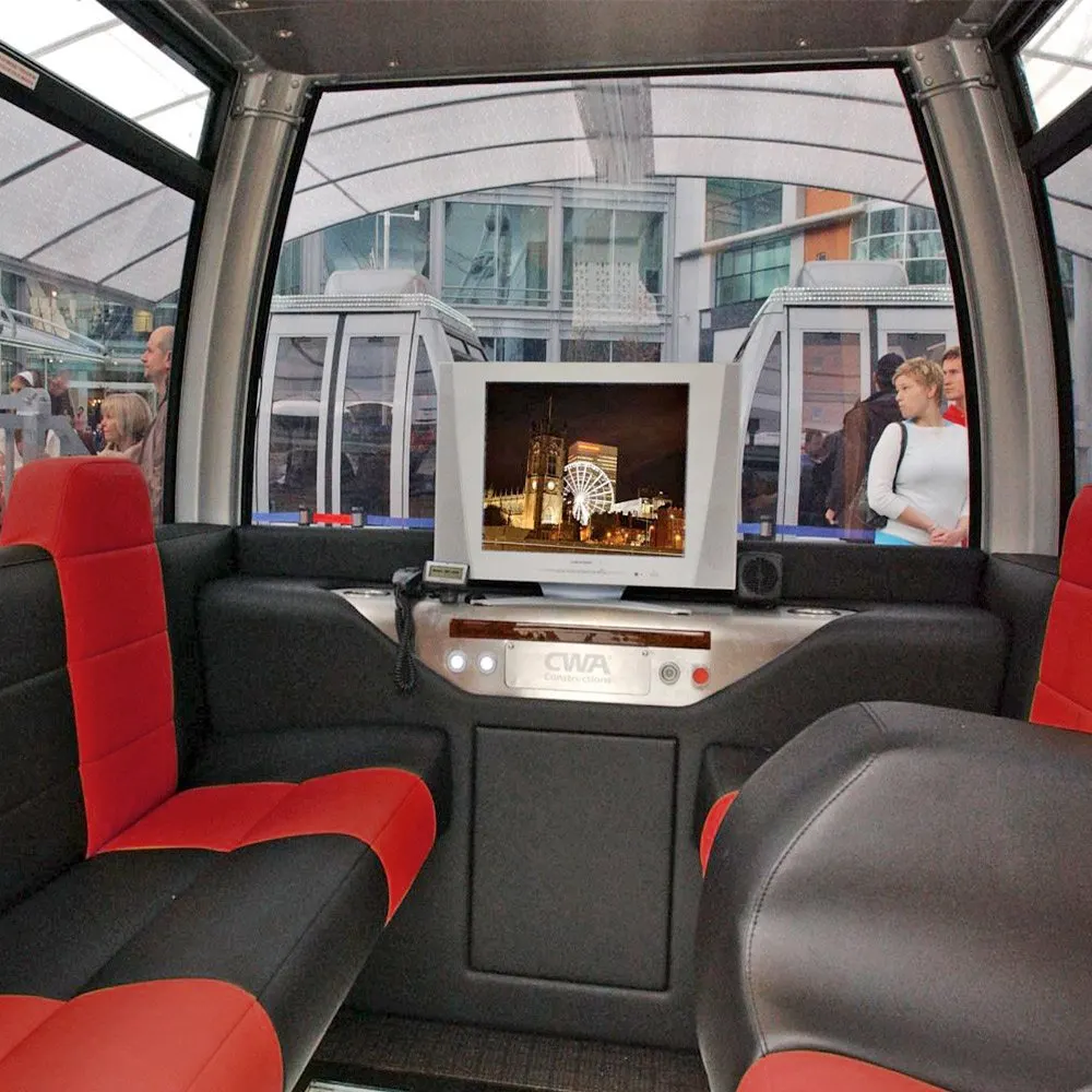 Interior of a passenger cabin on the London Eye with red and black seats, a screen displaying an illuminated cityscape, and visitors visible through windows.