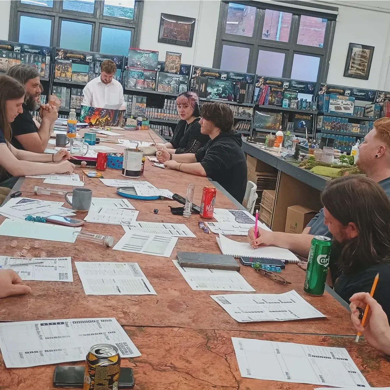 Group of people seated around a wooden table playing a tabletop game in a gaming shop with shelves of board games and miniatures in the background.