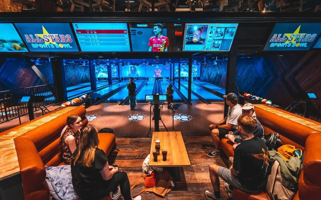 Groups of people bowling in a modern alley with illuminated lanes, orange seating areas, and digital scoreboards displaying game information.
