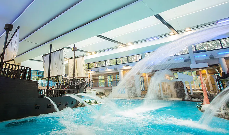 Water cascades down into a bright turquoise indoor pool beneath a modern ceiling, with a themed ship structure and viewing platforms visible.