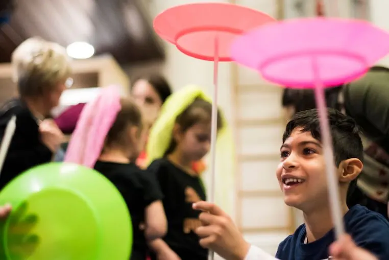 Young boy holding a colourful umbrella indoors with other children and adults at an indoor activity event.
