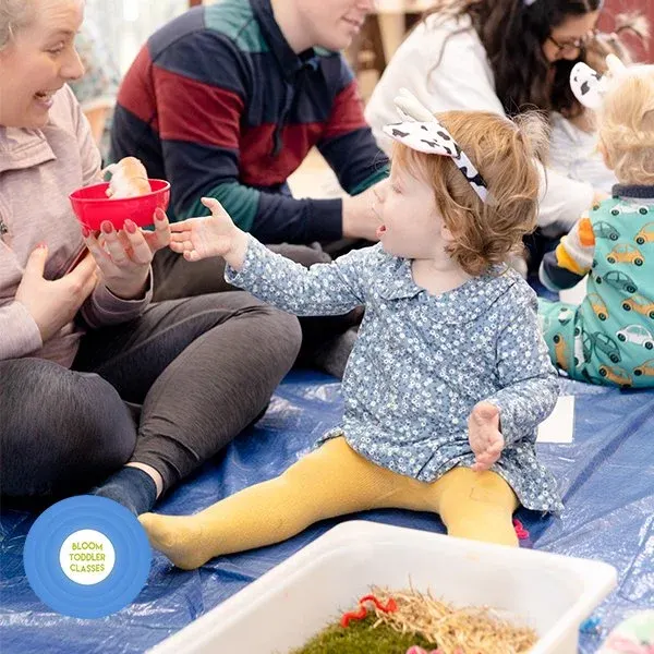 Young children and adults sitting on a blue mat during a sensory activity session with food and play materials.