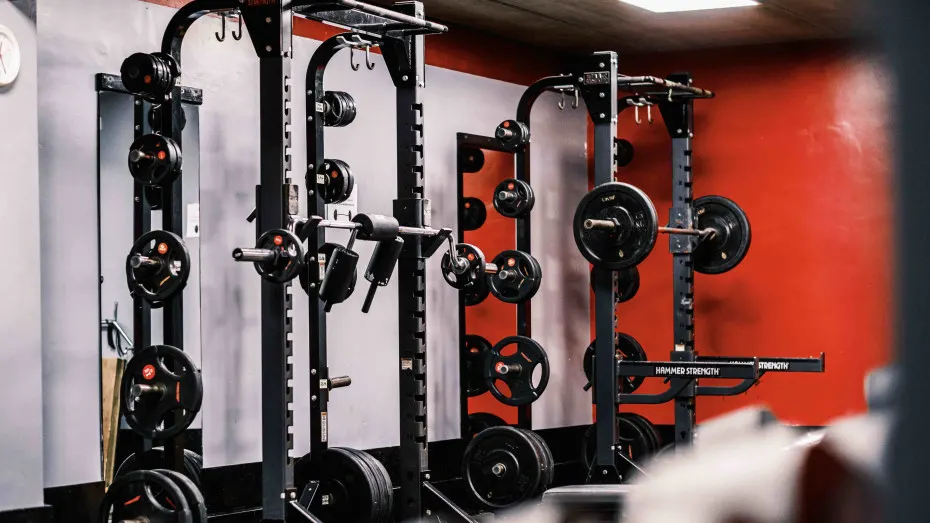 Multiple weight-training racks with barbells and weight plates in a modern gym with red and white walls.
