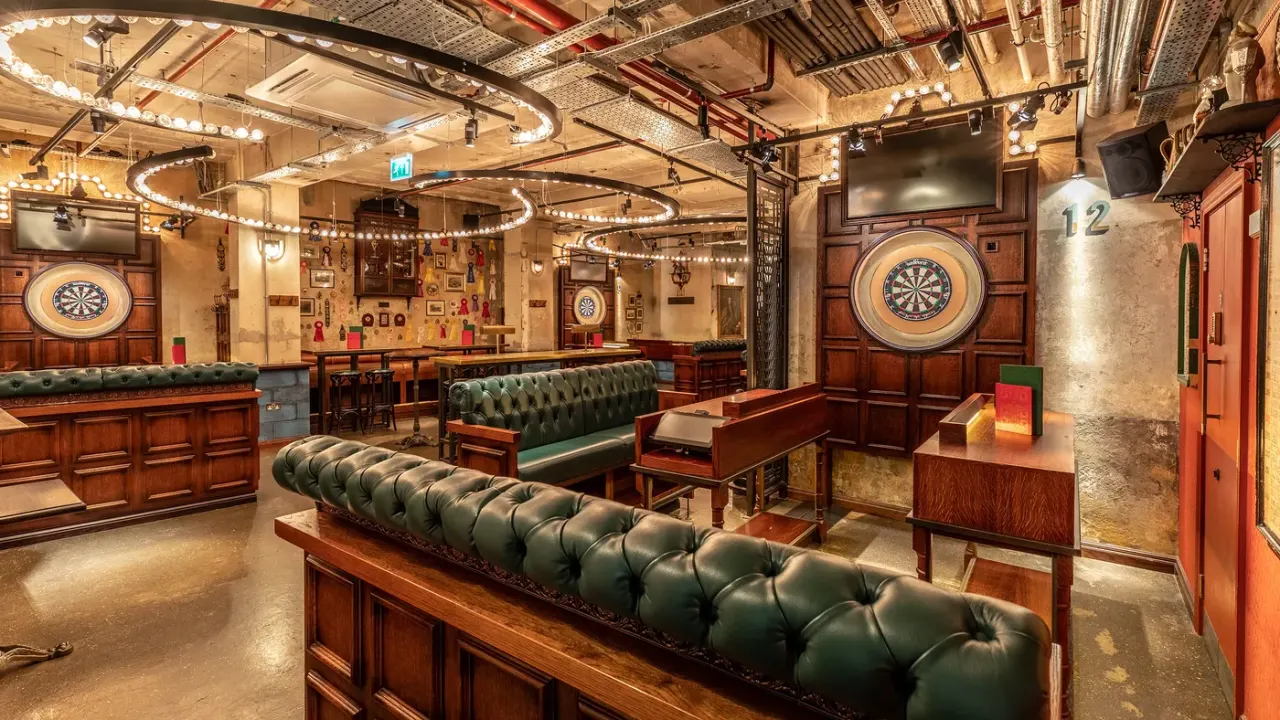 Upmarket darts bar with wooden panelling, green leather seating, illuminated dart boards, and decorative lighting overhead.
