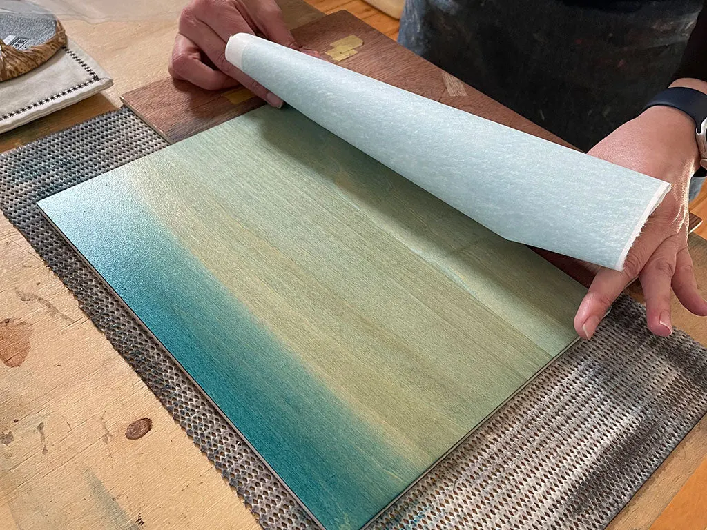 Person examining a roll of pale green and blue gradient fabric or wallpaper sample on a wooden table.