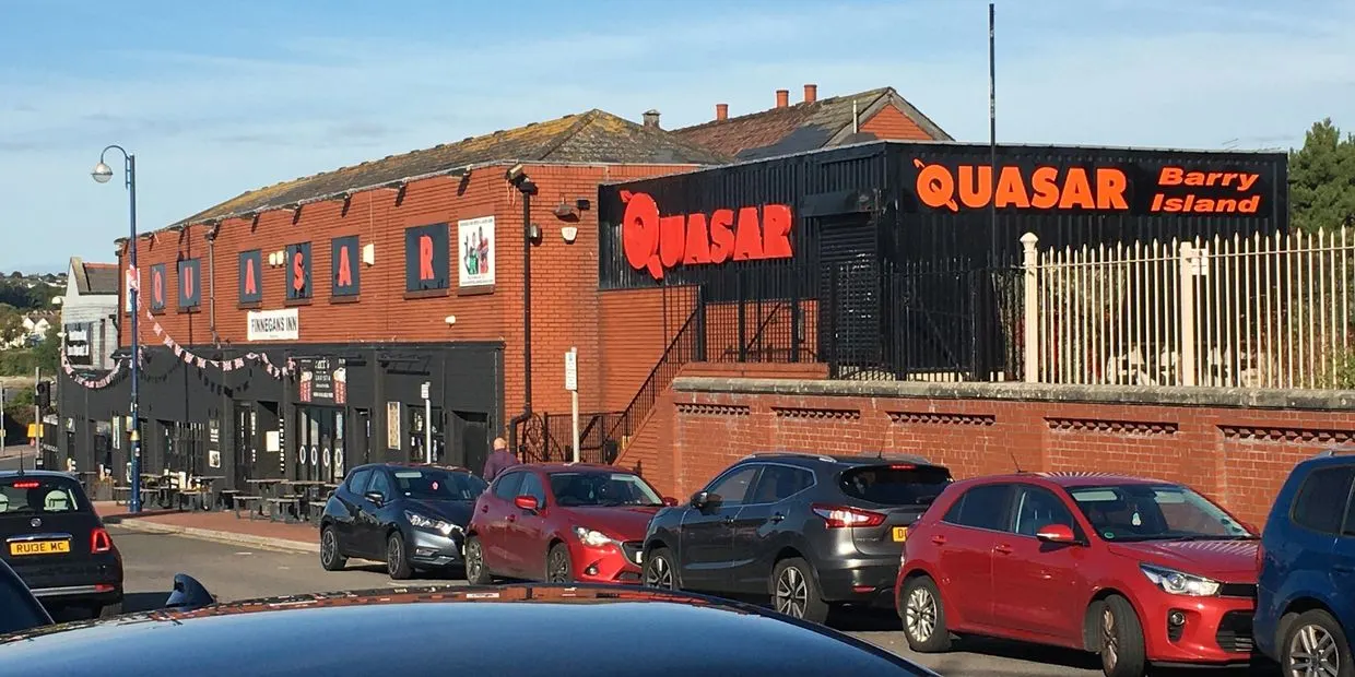 Red-brick leisure venue with "Quasar Barry Island" signage, parked cars in front, and residential buildings behind under blue sky.