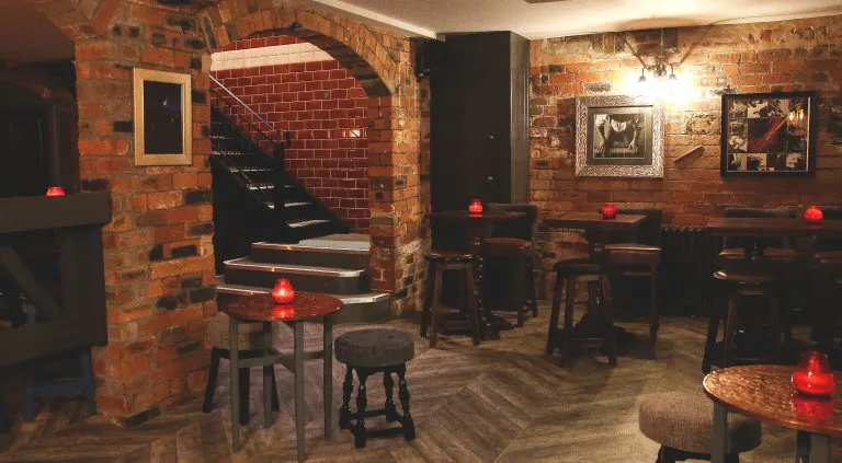 Interior of a basement bar with exposed red brick walls, wooden floors, bar stools, and red ambient lighting throughout the space.