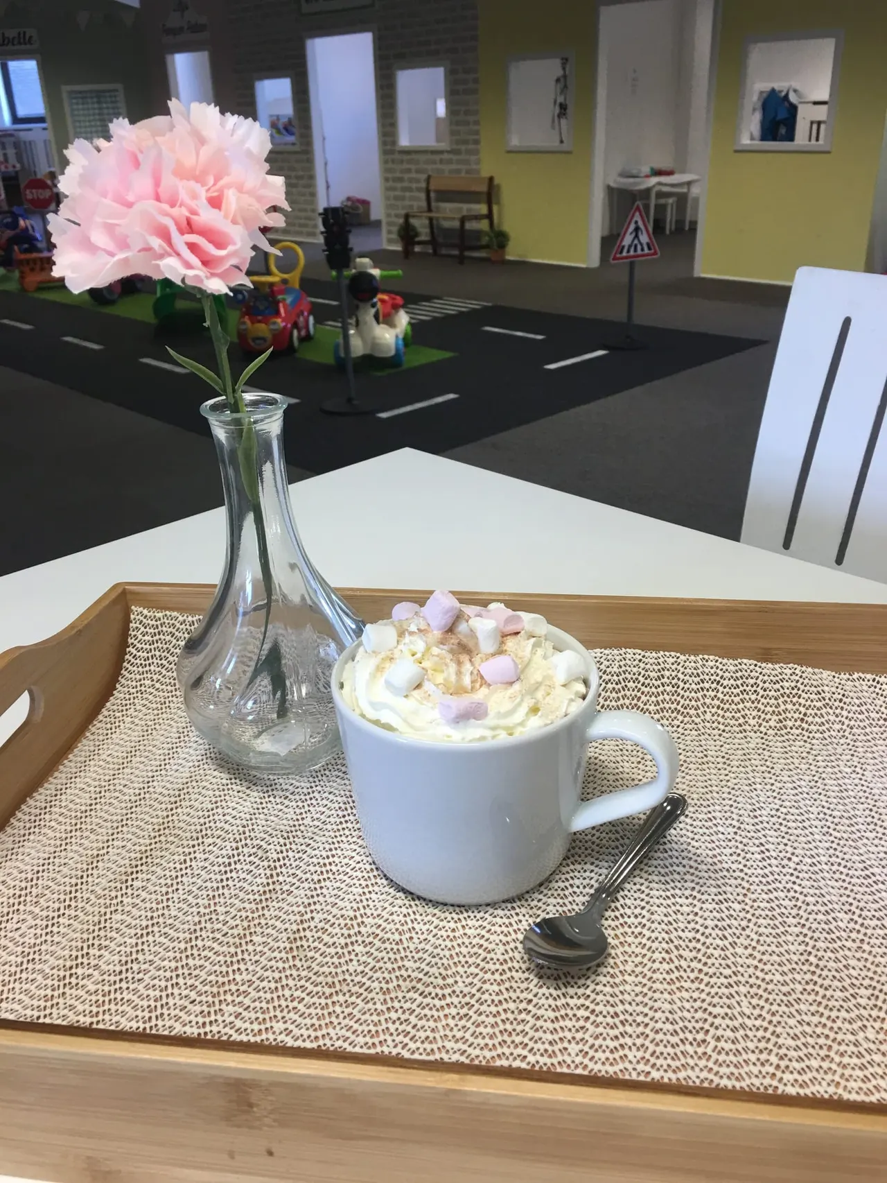 A white mug of hot chocolate topped with cream and marshmallows sits on a table beside a pink flower in a glass vase, in an indoor play area.