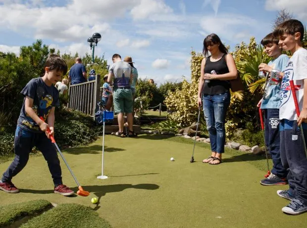 Children playing mini golf on a putting green with spectators watching on a sunny day.