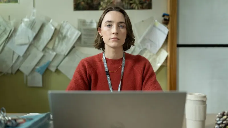 Person wearing a lanyard sits at a laptop in an office or workspace with papers pinned on the wall behind them.