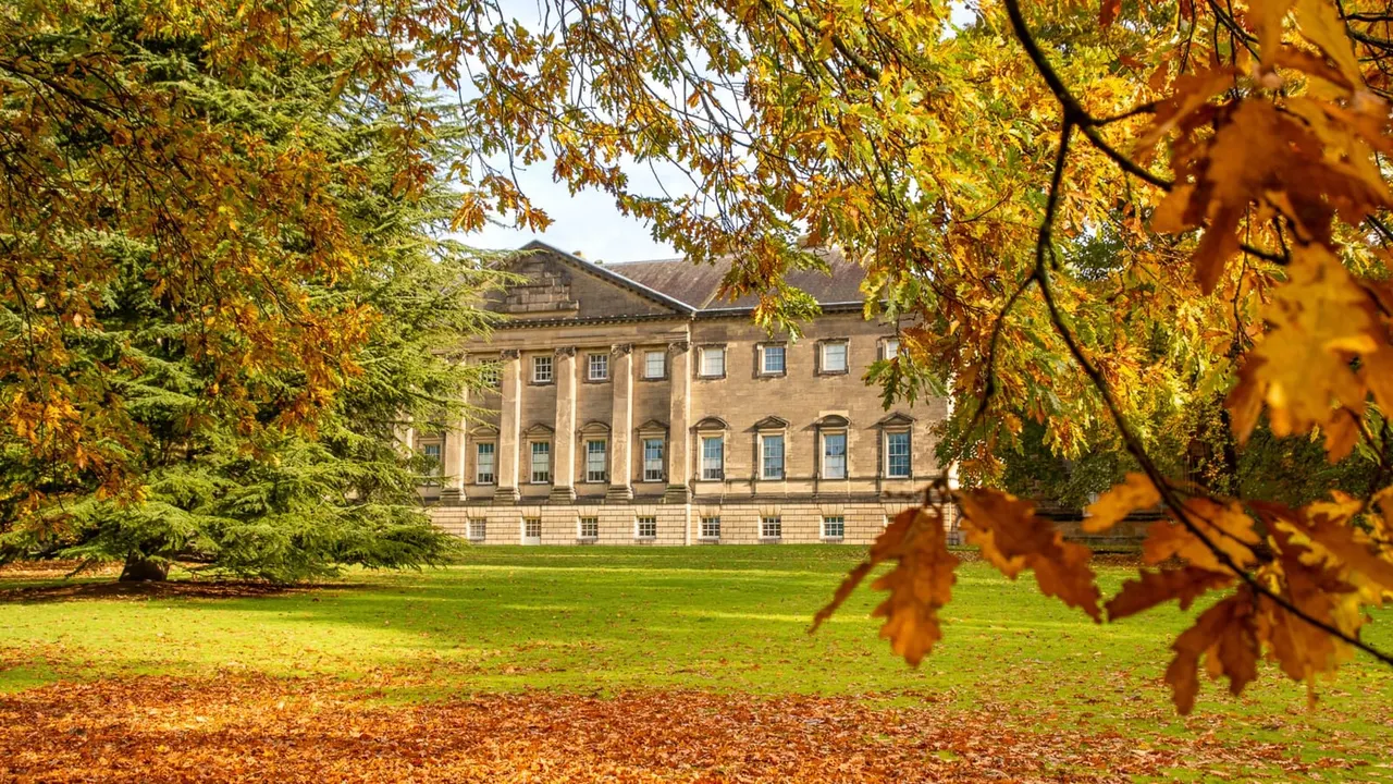 Autumn foliage frames a grand neoclassical stone mansion with columned portico overlooking manicured lawns.
