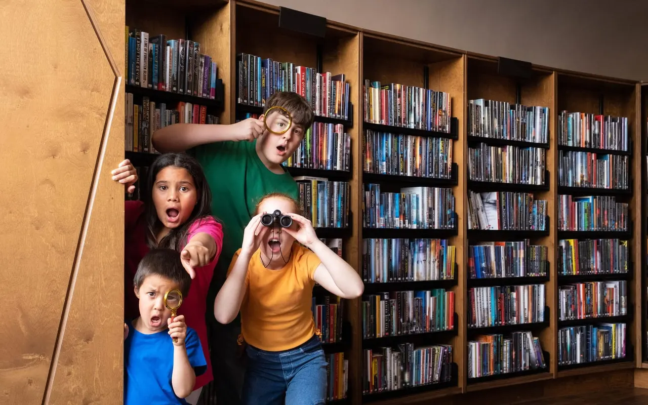 Four children posing playfully in front of wooden shelves stacked with DVDs and films, holding magnifying glasses and binoculars.