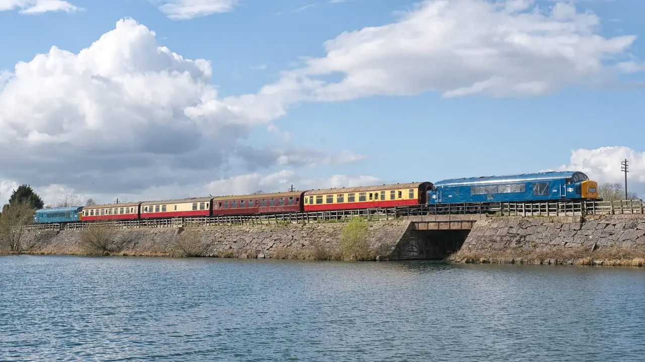 Heritage diesel train with red and yellow carriages crossing a bridge over water on a sunny day.