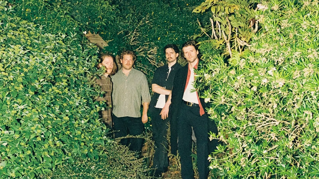 Four men standing in a narrow garden pathway lined with dense green foliage on both sides.