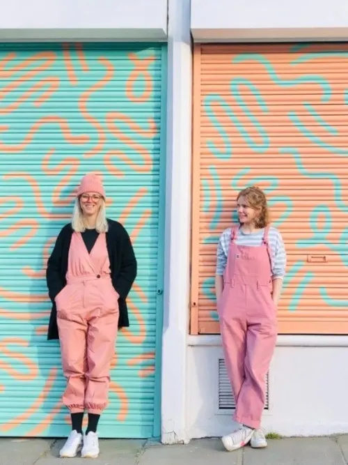 Two people in pink dungarees stand in front of colourful turquoise and orange striped shutters with geometric patterns.