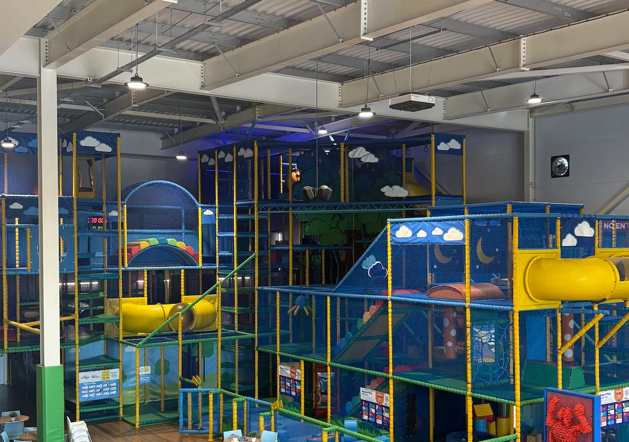 Multi-level indoor play structure with blue netting, yellow tubes and railings, and cloud-themed decorations in an industrial warehouse setting.
