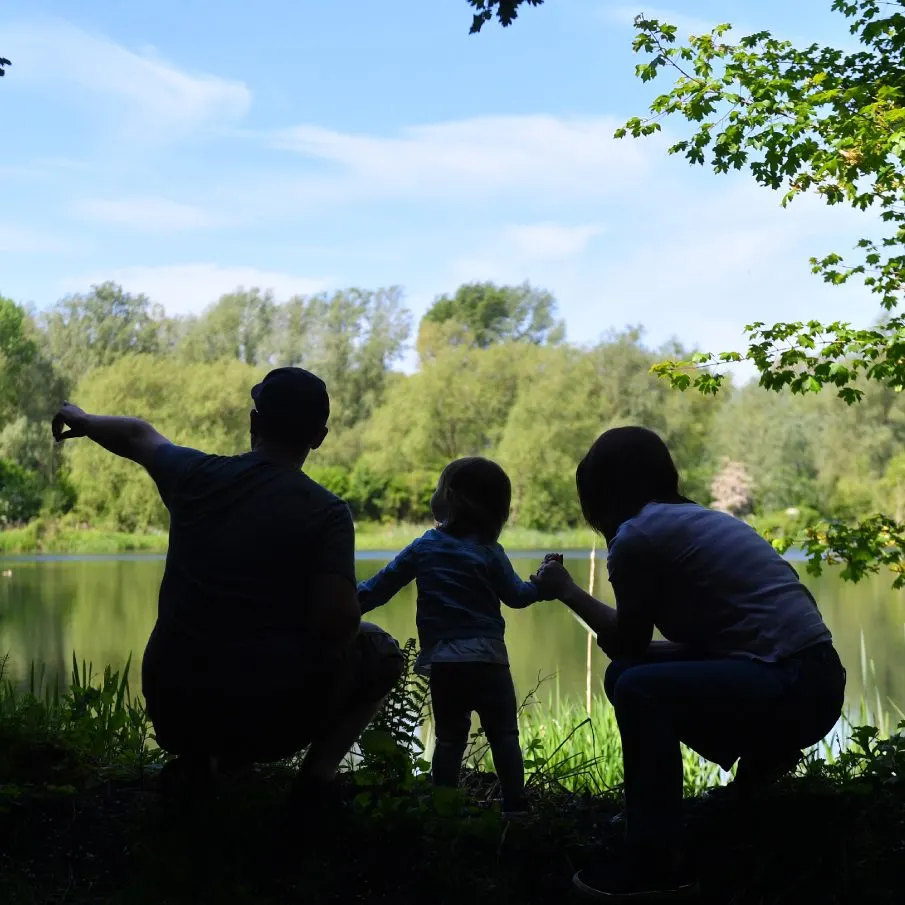 Adult and child silhouetted by a lake, surrounded by trees and greenery on a sunny day.