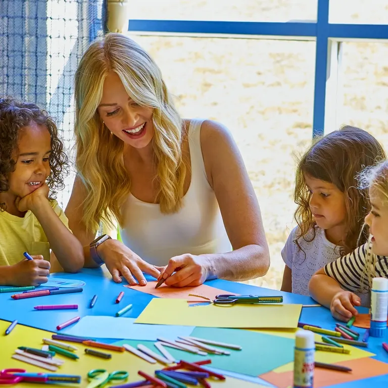 A woman leads a creative drawing activity with young children at a table surrounded by coloured markers and paper.