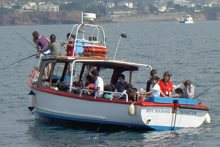 A red and white fishing boat filled with passengers on a coastal excursion, with cliffs and buildings visible in the background.