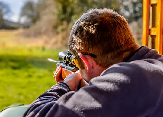 Person aiming a rifle with telescopic sight at an outdoor shooting range.