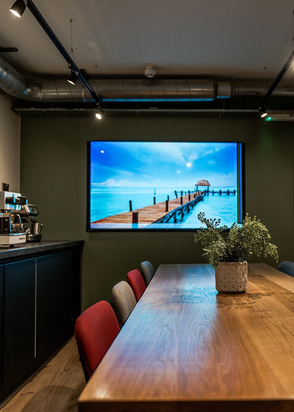 Modern hospitality lounge with wooden table, coloured chairs, and large screen displaying tropical beach pier scene.