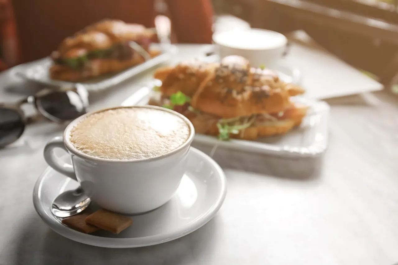 A cappuccino with latte art sits on a white table alongside plates of sandwiches and other food items at a café.