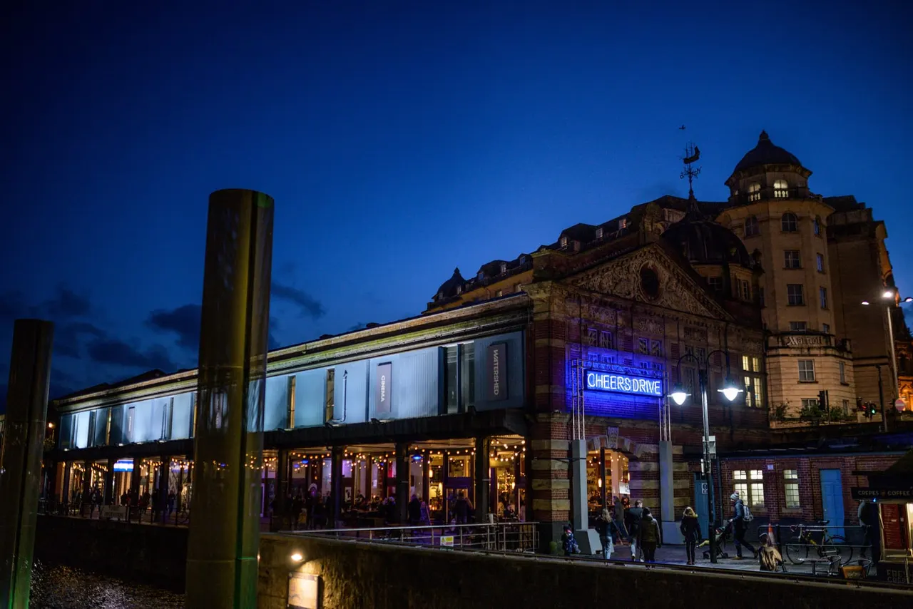 Waterfront venue building illuminated at dusk with blue neon signage, historic architecture visible beyond, people gathering outside.