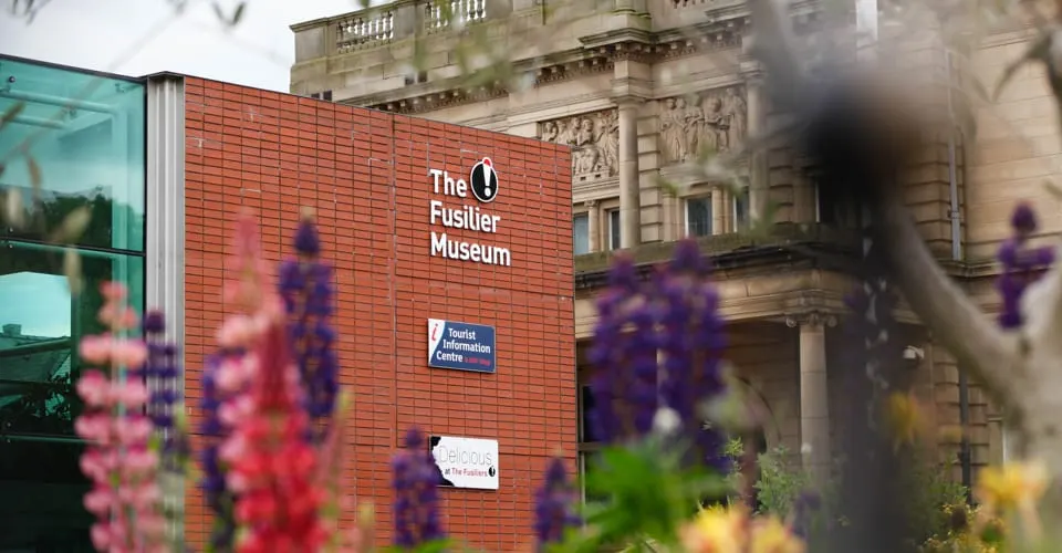 Red-brick museum building with classical stone architecture visible behind, viewed through flowering plants in foreground.