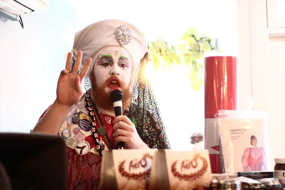 A performer in elaborate costume and makeup holds a microphone while speaking at a table during an event.