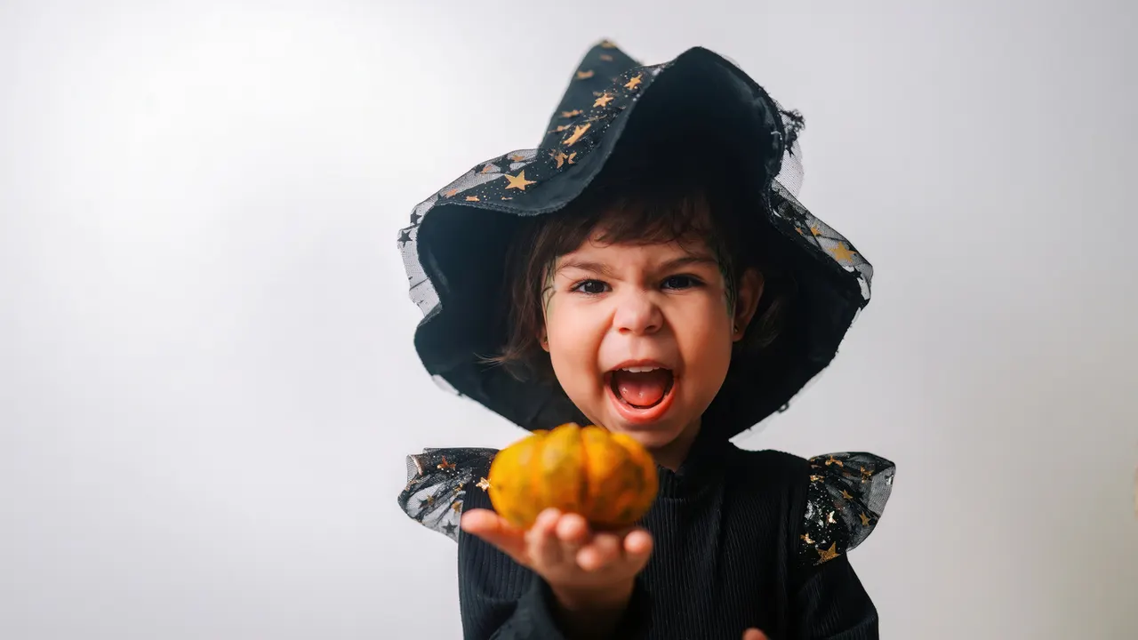 Child in black witch costume with star-patterned hat holding out a small pumpkin against white background.