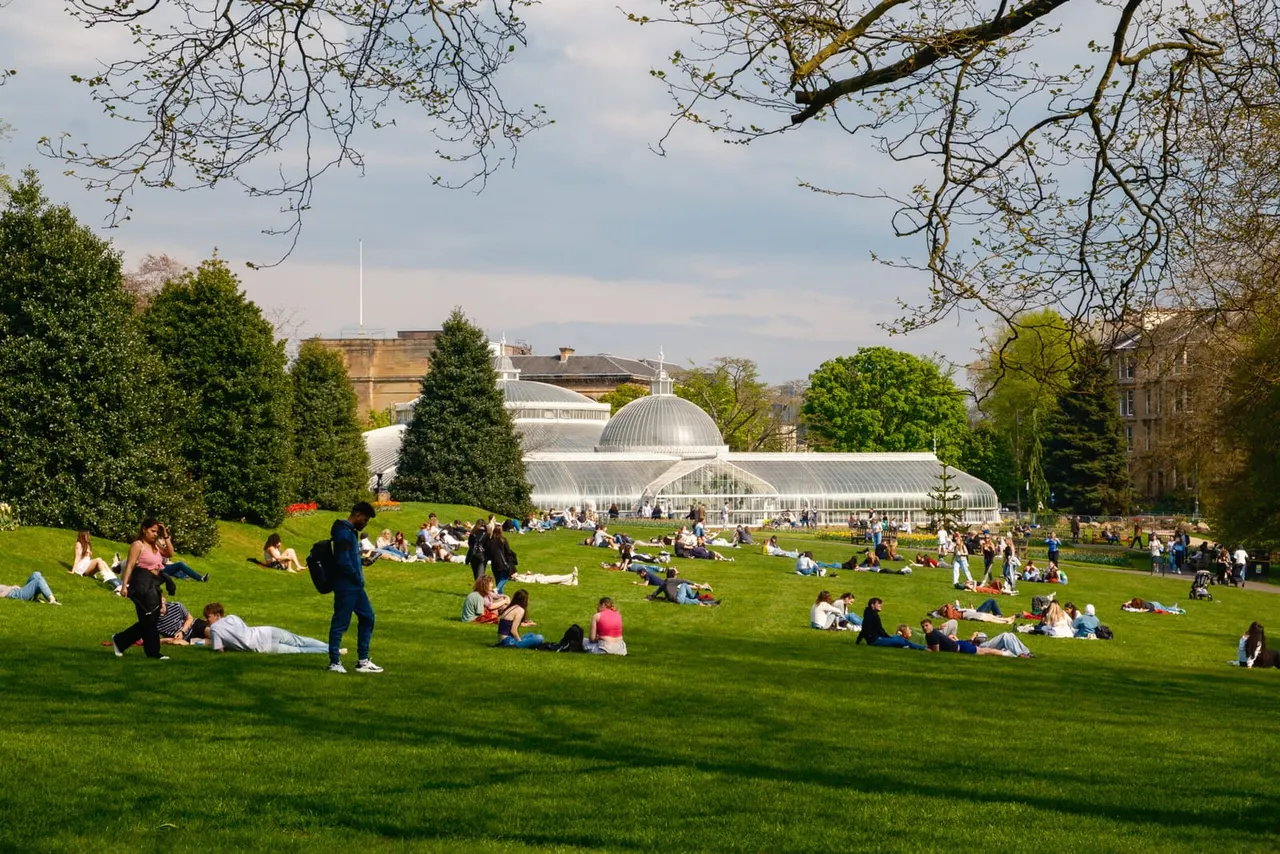 Crowds of visitors relax on a sprawling green lawn with a Victorian glasshouse and surrounding buildings visible in the background.