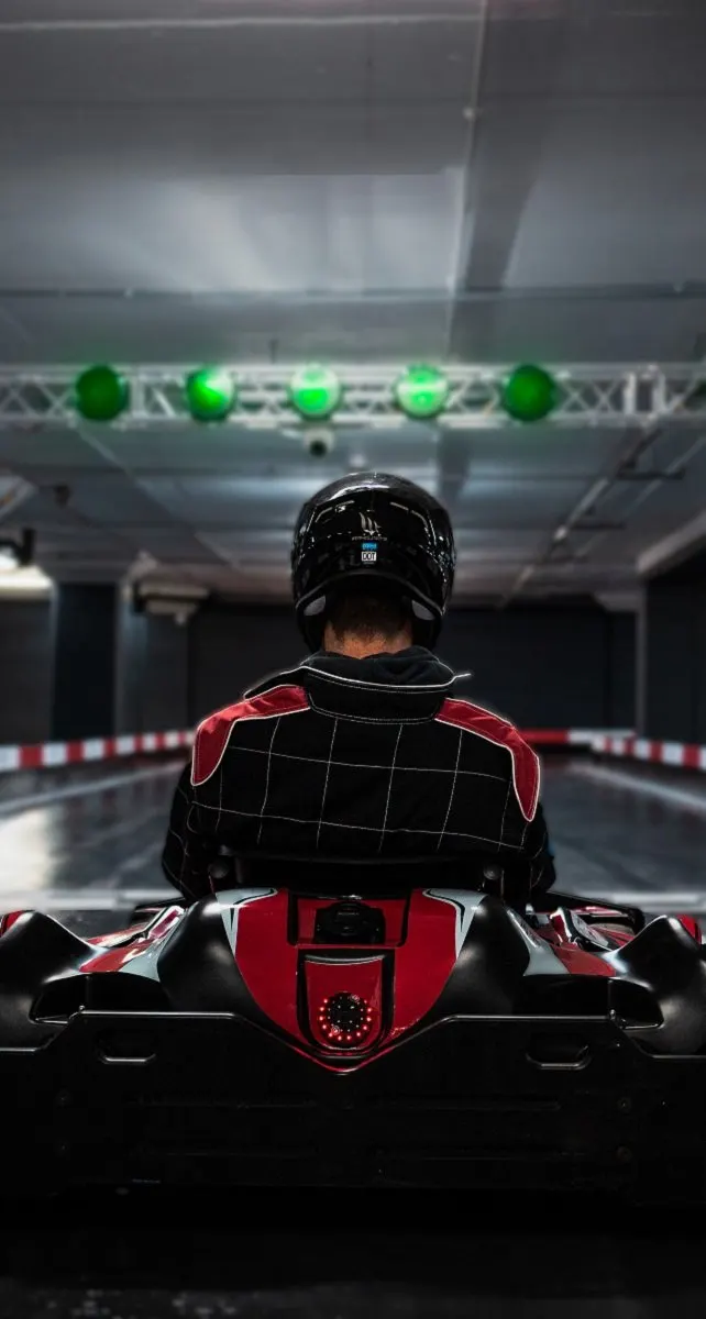 A racer in a black and red go-kart wearing a helmet, facing away toward a lit indoor track with green signal lights overhead.