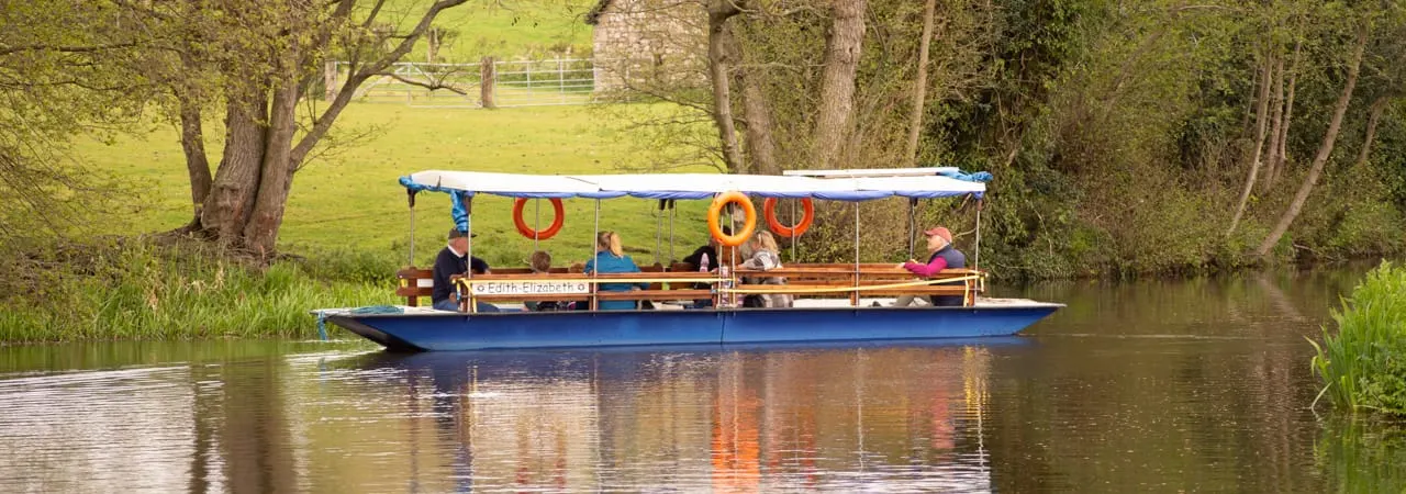 A blue and wooden canal boat with canopy carries passengers on a peaceful river lined with overhanging trees.