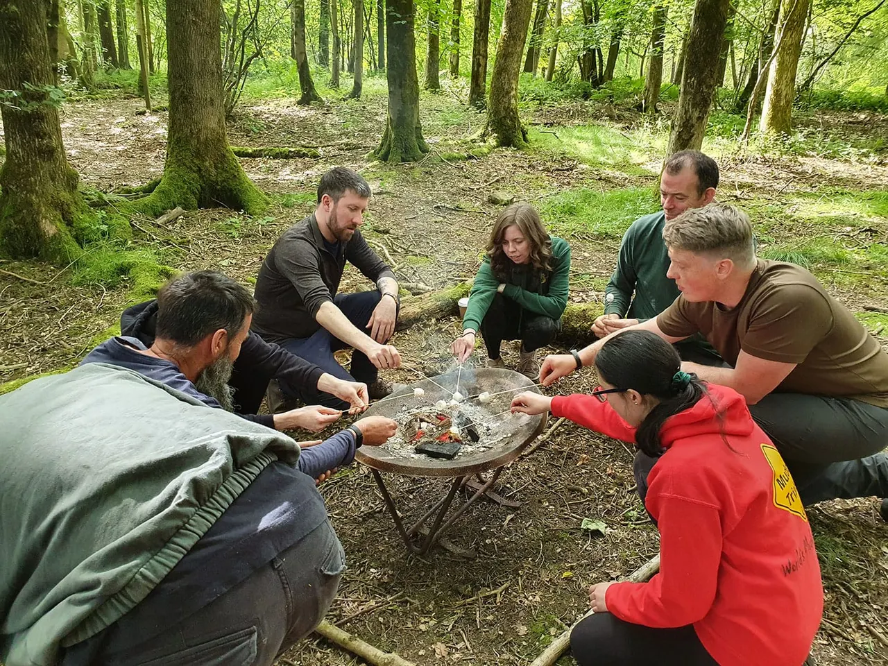 A group of six people gathered around a campfire in a woodland clearing, learning bushcraft skills.