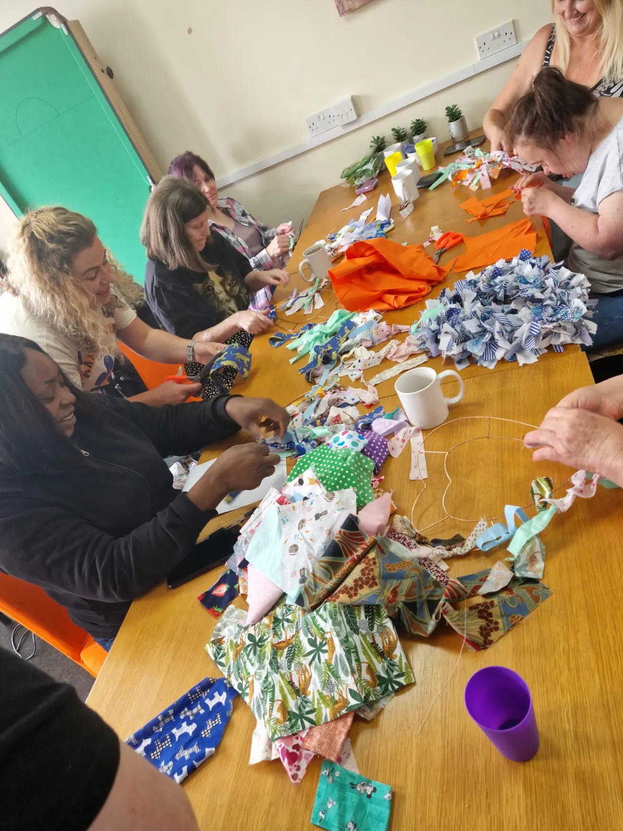 Group of adults and children gathered around a wooden table making colourful fabric crafts together in a community centre.