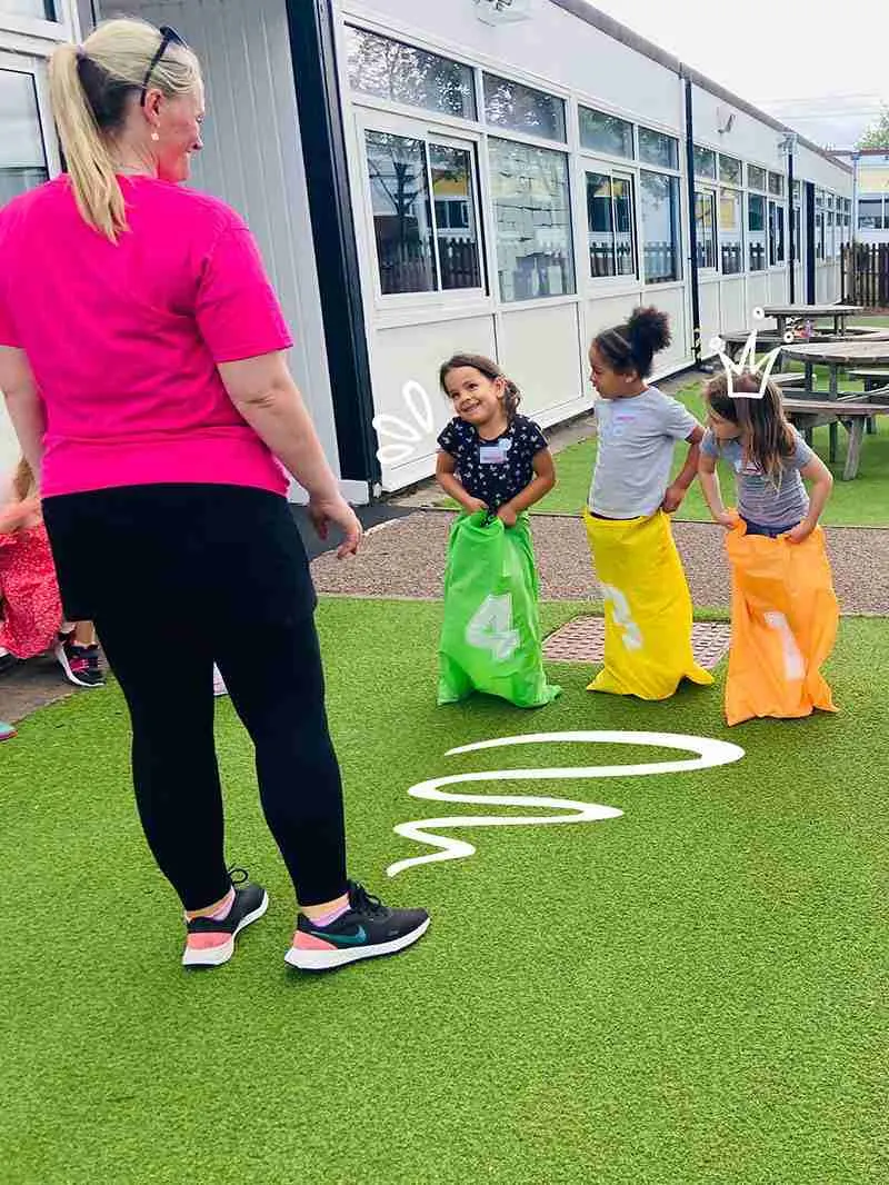 Adult supervisor leads children in a sack race activity on artificial turf outside a leisure facility.