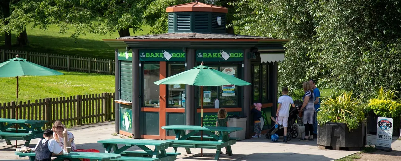 Visitors queue at The Bake Box café kiosk surrounded by green picnic tables and umbrellas in a sunny park setting with grass and trees.
