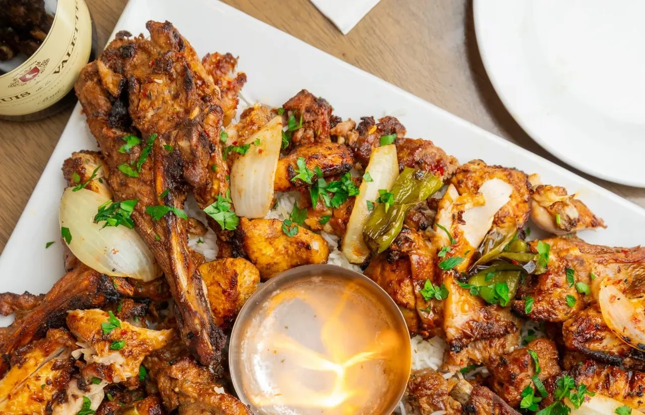 Grilled meat skewers with charred onions and peppers, served with dipping sauce on a white plate.
