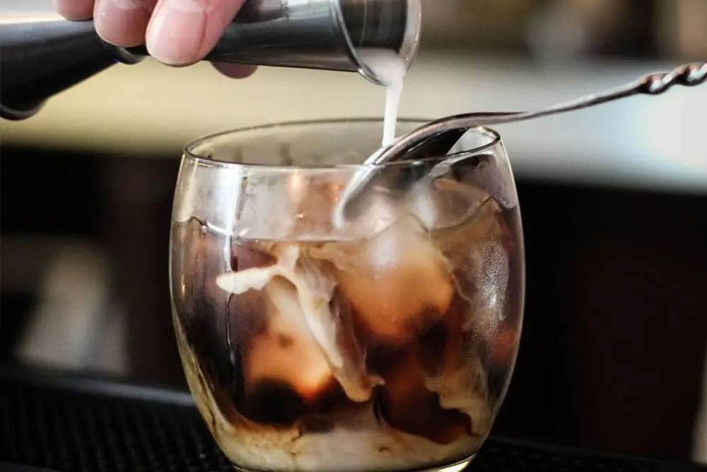 Bartender pouring cream into a glass of dark iced cocktail.