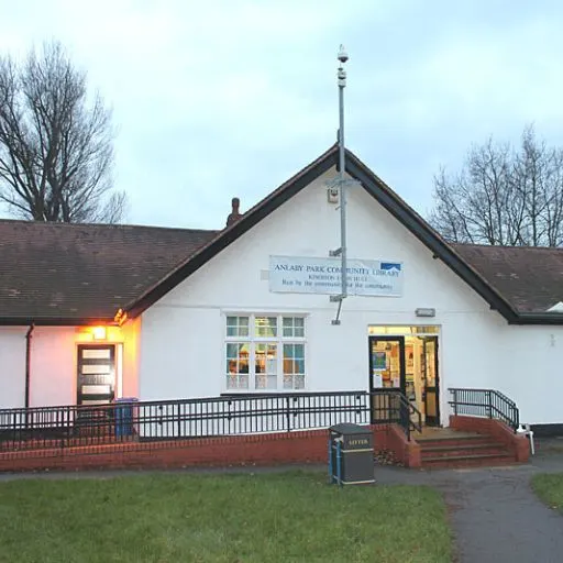 White-painted community library building with brick base, lit entrance doors, and bare trees in background at dusk.