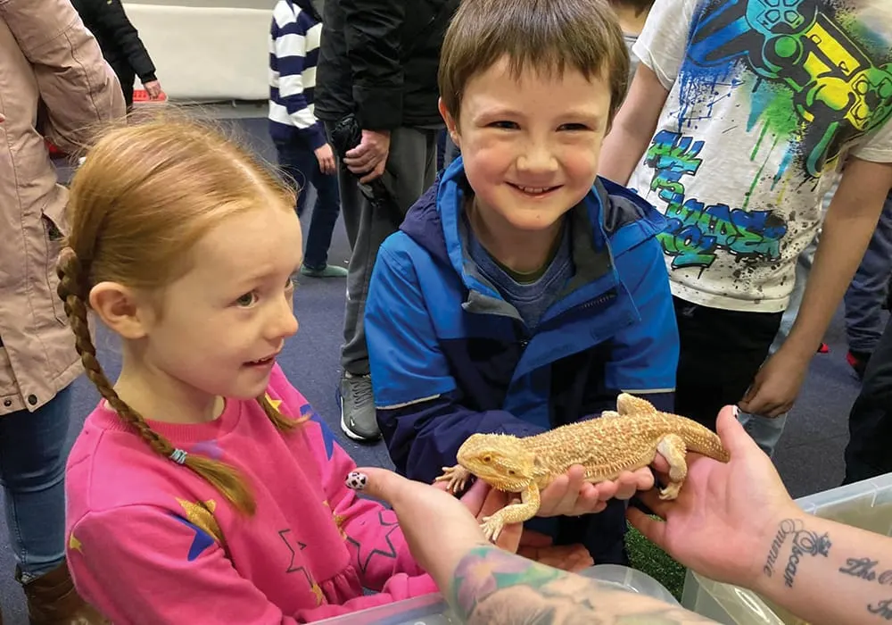Two children smiling while holding a bearded dragon during a hands-on animal encounter with other visitors watching.