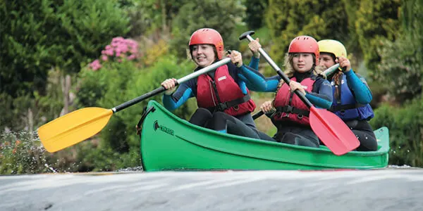 Three people in helmets and life jackets paddling a green canoe on a river with trees behind.