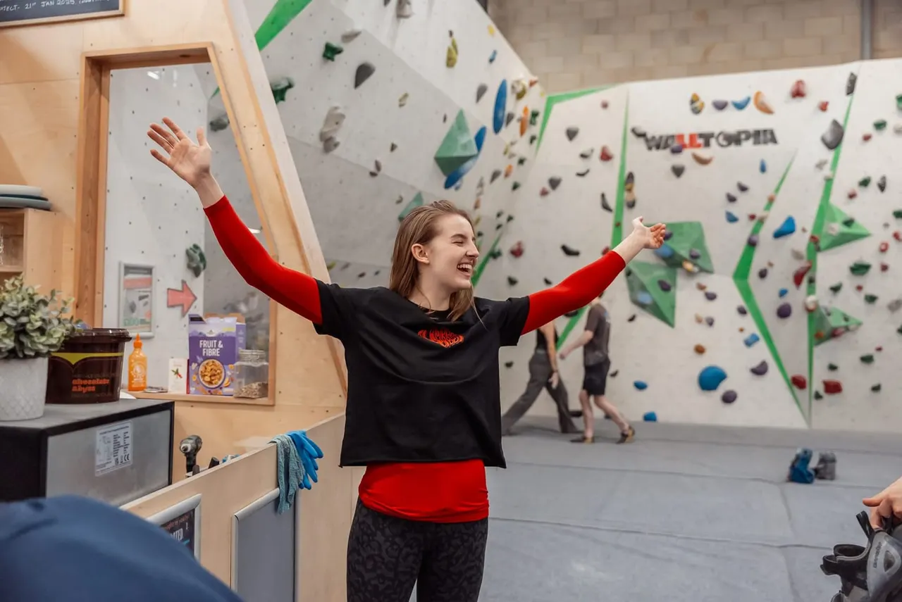 Young person celebrating with arms raised in front of indoor rock climbing walls with coloured holds.
