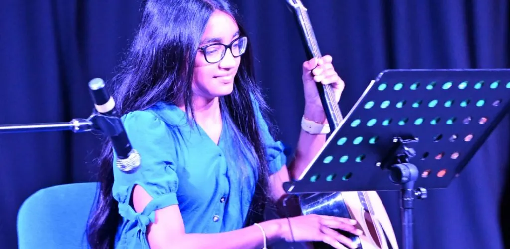 Young musician playing bass guitar whilst reading sheet music on stage under blue lighting.