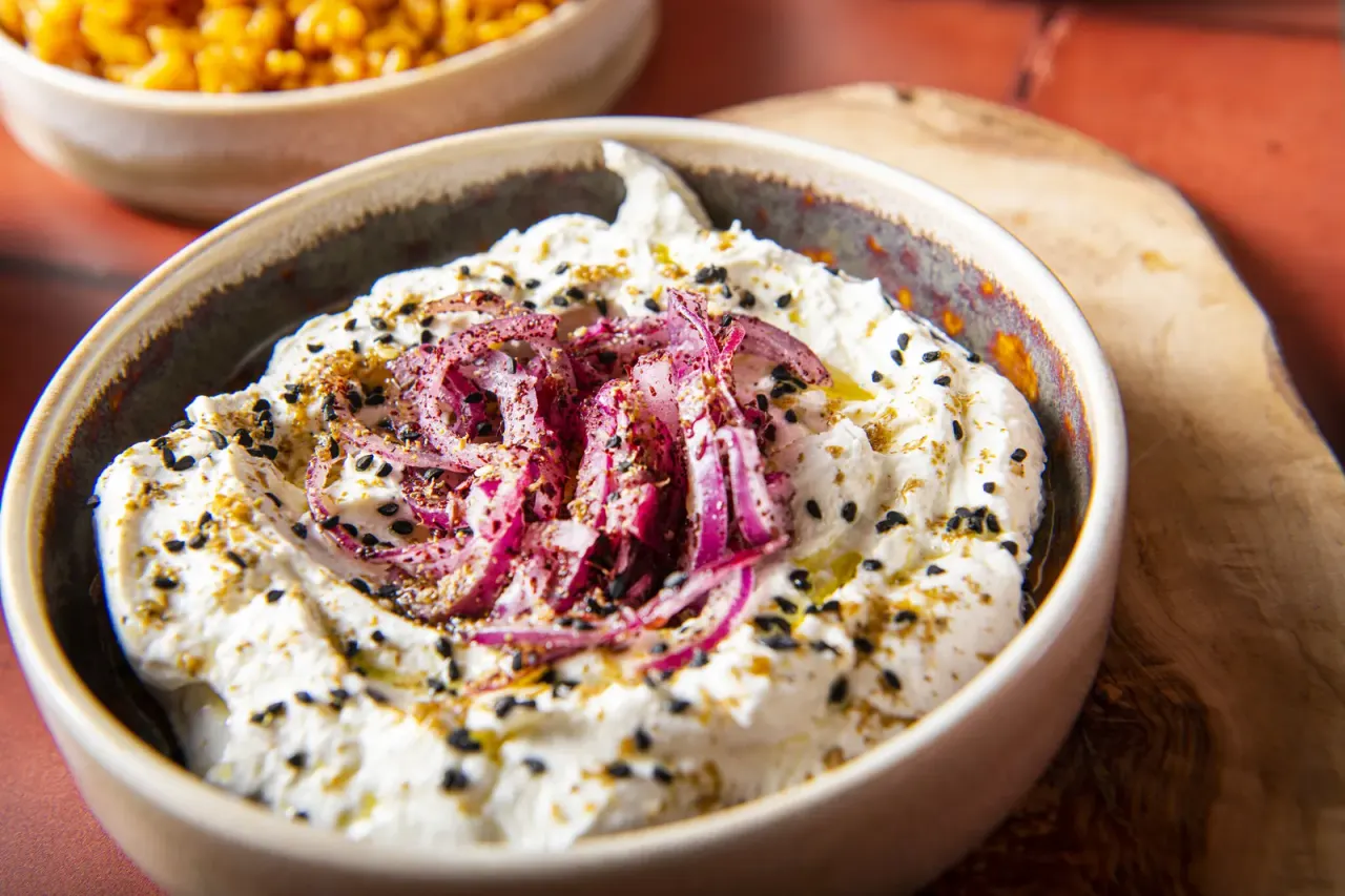 Bowl of hummus topped with roasted beetroot strips, black sesame seeds and olive oil, with corn in a dish behind.