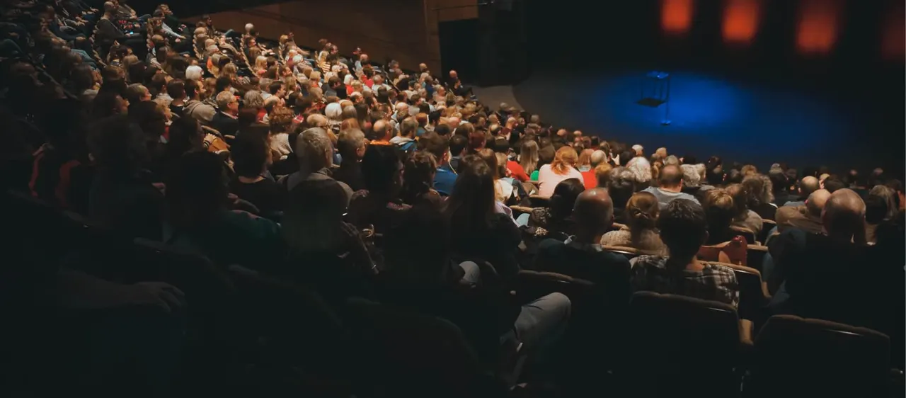 A crowded theatre auditorium with hundreds of audience members seated, watching a performance on a blue-lit stage.
