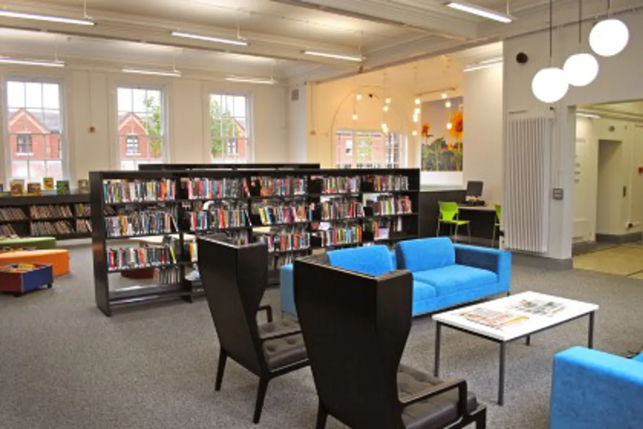 Modern library interior with bookshelves, blue seating, and large windows overlooking brick buildings outside.