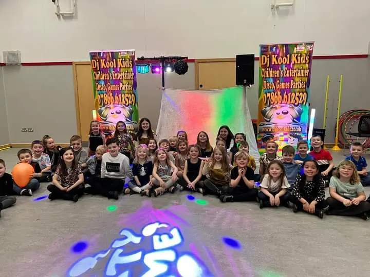 Large group of children seated on floor at a children's entertainment venue with DJ equipment and colourful stage lighting.