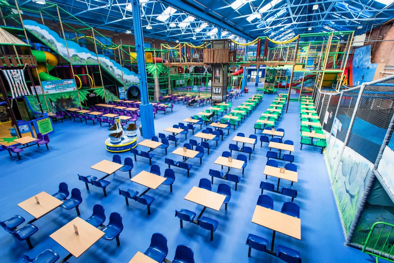 Interior of an indoor play centre with rows of picnic tables and benches, climbing structures, and soft play equipment beneath a blue roof.
