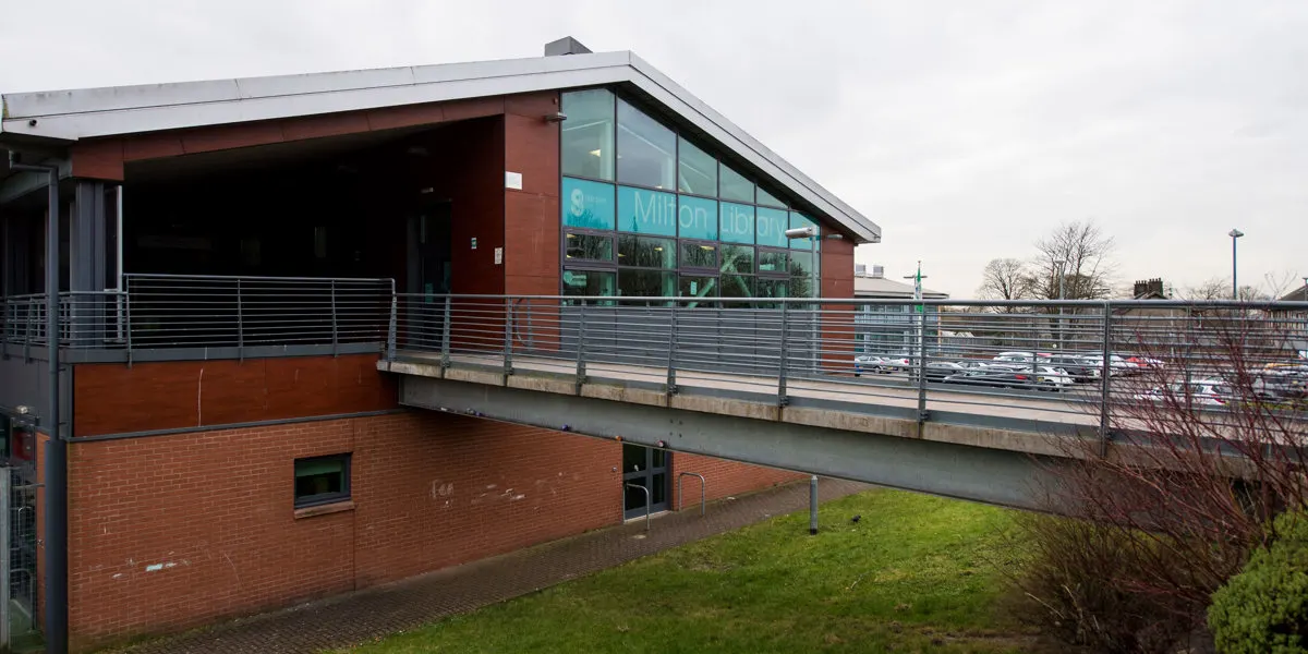 Modern brick and glass library building with elevated deck and metal railings, surrounded by grass and bare trees.